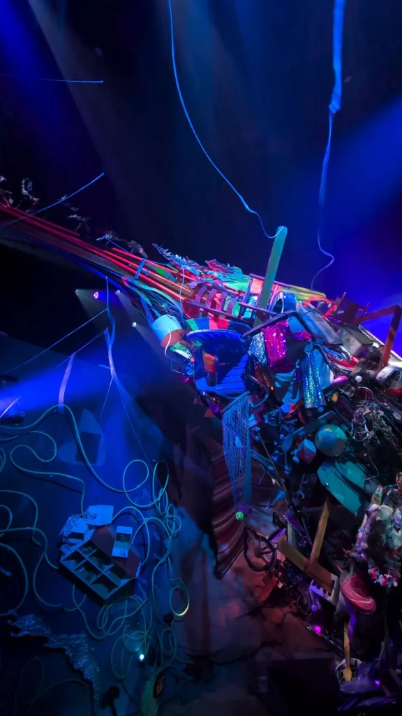 A very eclectic art and massive art sculpture comes in from the right. It's filled with many bright colors and strange objects. There's lights shining on it from the left. The rest of the room is covered in a calming blue glow.